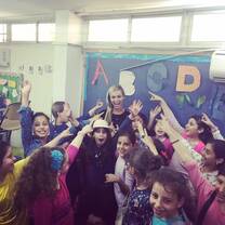 A smiling teacher stands in a classroom, surrounded by smiling students pointing at her