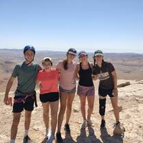 5 teens with their arms around each other stand on top of a mountain after a hike.