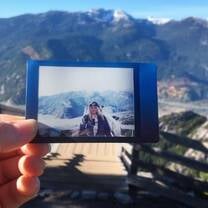 a girl holds out a polaroid of herself in front of a mountain, with the mountain range in the background