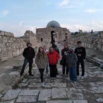 A group of 5 students stand in a row on an ancient paved road with ruins in the background.