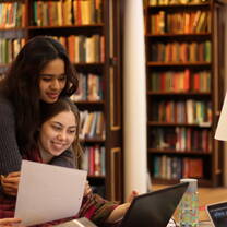 Two students hug while looking a computer in Lady Margaret Hall College Library