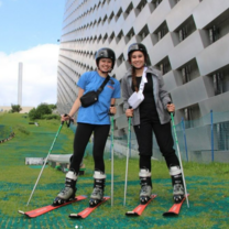Two students are standing on an artificial grass slope and are wearing skis