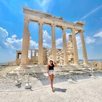 A young woman with long brown hair, wearing a black tank top and light denim shorts with a straw hat, stands in front of temple ruins with columns still standing.