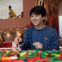 A male student is smiling and sitting at a table. The table is covered with lego bricks. In the background there is a dinosaur built with lego. 