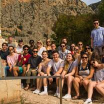 A group of students are sitting on some rocky steps and smiling for the camera. They are dressed in summer clothing and many have sunglasses on. Behind them is a rocky cliff face and stone wall.