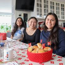 Three girls are sitting at a table and smiling. In front of them is a basket of baked good and Danish flags.