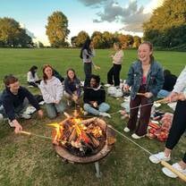 A group of young students are sitting around a fireplace and cooking bread on long sticks.