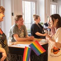 Two students are chatting to people sitting at a table that says Queer the Future. There are rainbow flags on the table.