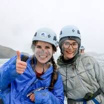 Two girls are wearing helmets and warm clothing. They are smiling and one is giving a thumbs up. The background is a glacier.