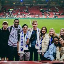 A group of students are standing in front of the pitch of a football stadium. They are wearing FC Copenhagen blue and white scarves.