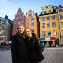 Two female students are standing in an old town square with colorful historical buildings behind them.