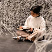 A girl is sitting cross legged on the floor, writing in a book. She is surrounded by white string in a webbed pattern from floor to ceiling.