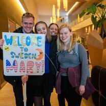 A mother, father and their daughter are standing with a newly arrived student. The family is holding a homemade sign that says 'Welcome to Sweden Mary' 