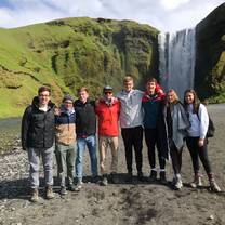 A group of young people are standing in front of a waterfall. A lush green cliff is also behind them.