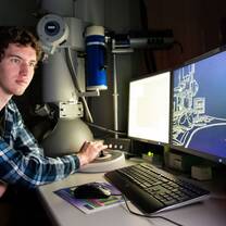 A male student is sitting in front of a machine and three computer screens. 