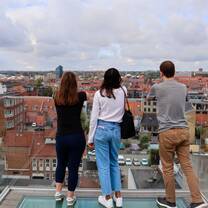 Two young women and a man are standing on a balcony and looking out at a city scape. Their backs are to the camera.