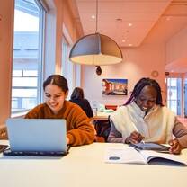 Two female students are sitting at a table studying. One is looking at a laptop screen while the other has books open in front of her.
