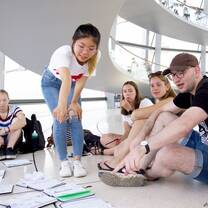 A female student is standing and pointing at some papers on the ground. Other students are sitting on the floor also looking at the papers.