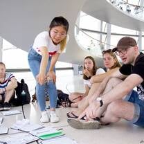 A female student is standing and pointing at some papers on the ground. Other students are sitting on the floor also looking at the papers.