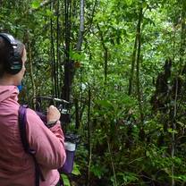 a girl in pink jacket holds audio recording equipment in dense forest