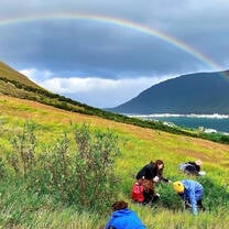 Students on a grassy hill, a rainbow overhead and a hill in the distance.