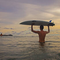 Boy holds surfboard above his head as he treks through the calm ocean waters