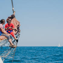 Two people sitting on the bowsprit of a schooner.