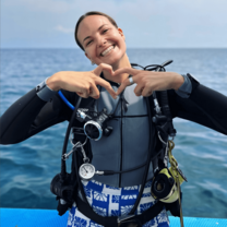 A happy project laut intern sitting on the edge of the dive boat in full gear smiling and making a heart with her hands
