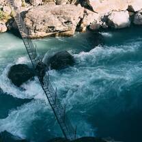 A NOLS student crosses a bridge over fast, blue water.