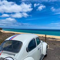 A classic white vintage car parked along Spain’s beautiful coastline with golden sands and turquoise waters under a bright blue sky, capturing the adventure and scenic beauty participants experience with RVF International's Teach English in Spain program.