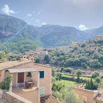 A serene hillside village in Spain with rustic stone homes, lush greenery, and towering mountains under a bright blue sky.