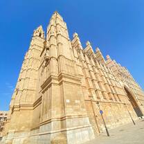 A stunning view of a towering Gothic-style cathedral with intricate stone details set against a clear, vibrant blue sky. The grand structure's spires and architectural elements showcase its historic beauty in a sunlit plaza.