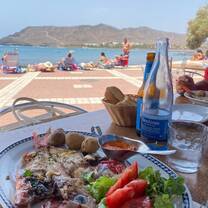 A plate of fresh grilled fish with roasted potatoes, salad, and tomatoes, served on a seaside table with a view of the beach, mountains, and clear blue water in Spain. A glass bottle of water, a beer, and a bread basket sit on the table, capturing a sunny coastal dining experience.