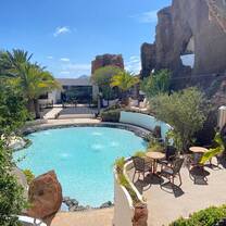 A tranquil pool surrounded by volcanic rock formations, lush greenery, and outdoor seating under a clear blue sky, showcasing the peaceful retreats and natural wonders experienced with RVF International’s Teach English in Spain program.