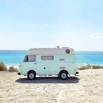 A pastel blue camper van parked on a sandy coastal road with the sparkling turquoise sea and a clear blue sky in the background, ready for a Spanish road trip adventure.