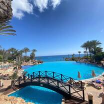A breathtaking resort pool in Spain with crystal-clear water, surrounded by sun loungers, palm trees, and parasols. A charming wooden bridge crosses part of the pool, with views stretching to the serene blue ocean under a cloudless sky. Guests enjoy the sunny atmosphere, showcasing the perfect Spanish coastal escape for RVF International participants exploring Spain.