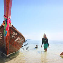 A longtail boat rests on white sand with crystal blue waters behind. Students are wearing snorkelling gear and wading in the water. 