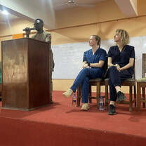 Two volunteers sit on a stage in scrubs, looking up at a man speaking at a podium