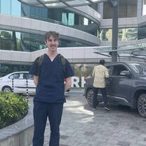 A volunteer wearing scrubs stands smiling in front of a hospital. Behind him, big white letters read 'I HEART RHL'