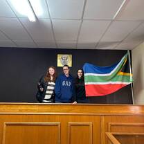 Three volunteers stand in a courtroom with the South African flag