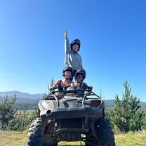 Two volunteers sit on a quad bike, with another volunteer behind them reaching into the blue sky