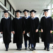 Five graduates in caps and gowns walk together down a hallway, facing forward and looking at the camera.