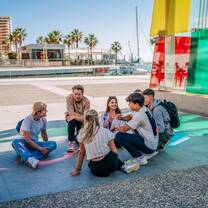 EF Gap Year students relaxing and chatting in the sunshine near the Pompidou Centre in Málaga, Spain, surrounded by palm trees and harbor views during their language and culture experience.