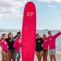 EF Gap Year students on the beach in Tamarindo, Costa Rica, posing with a bright pink EF surfboard after a surf lesson, smiling under the sun by the ocean.