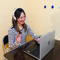 A Japanese teacher conducting an online 1-on-1 lesson with a student via Zoom. The teacher is smiling and explaining a lesson in Japanese using teaching materials on the screen.