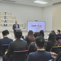 A man in a suit stands at a podium decorated with a blue floral arrangement, speaking in front of a screen displaying AIHM College and NCUK Study Centre logos in a classroom setting.