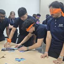A group of students wearing dark blue shirts work together on a puzzle. Two students are blindfolded with orange bandanas, while others watch and assist. They are gathered around a light-colored table in a classroom.