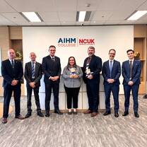 Seven people in business attire stand in a row, smiling, inside an office with the AIHM College and NCUK logos on the wall behind them. The woman in the center holds two stuffed animal toys.