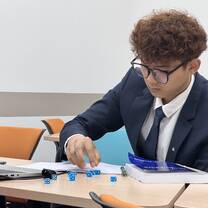 A young man in a suit and glasses sits at a classroom desk, arranging blue dice on papers beside a laptop and a large book, focused on his work. Other students are in the background.