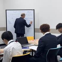 A teacher in a suit writes on a whiteboard labeled Simple Probability, while three students in suits sit at desks, taking notes and using laptops in a classroom.